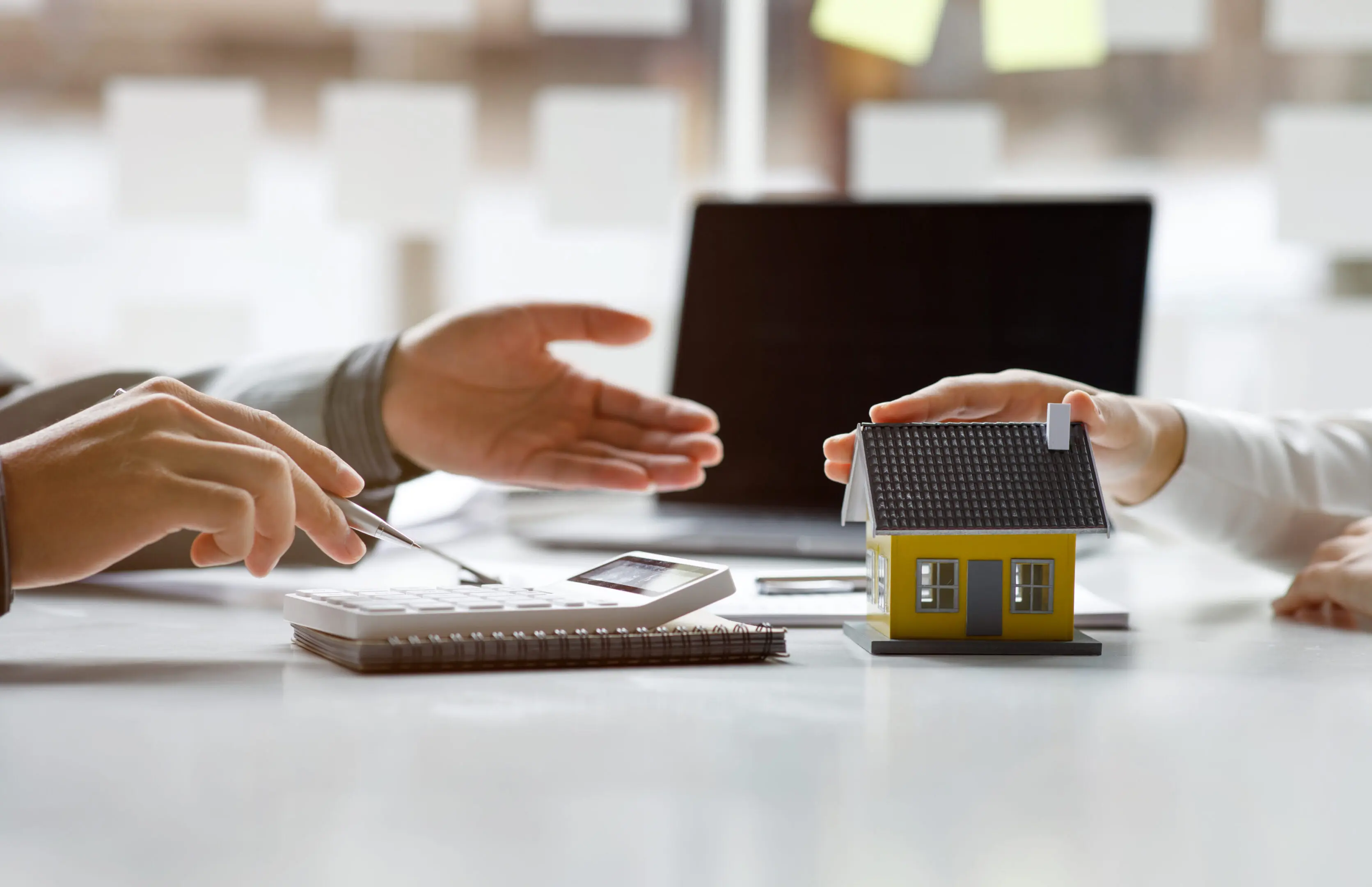 Two people discuss documents at a desk with a calculator, notepad, and a small model house, with a laptop in the background.