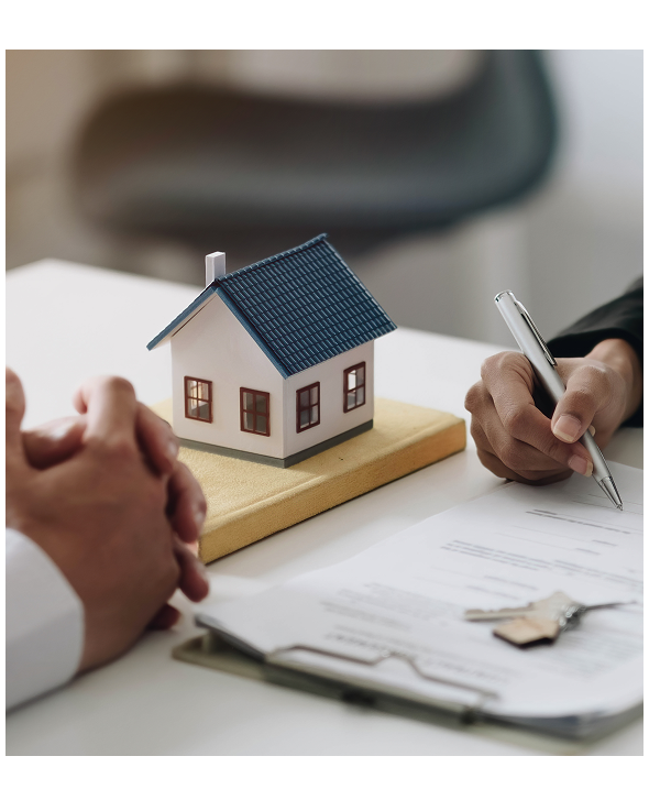 Two people sit at a desk with a model house, contract papers, and keys, suggesting a real estate or home purchase agreement.