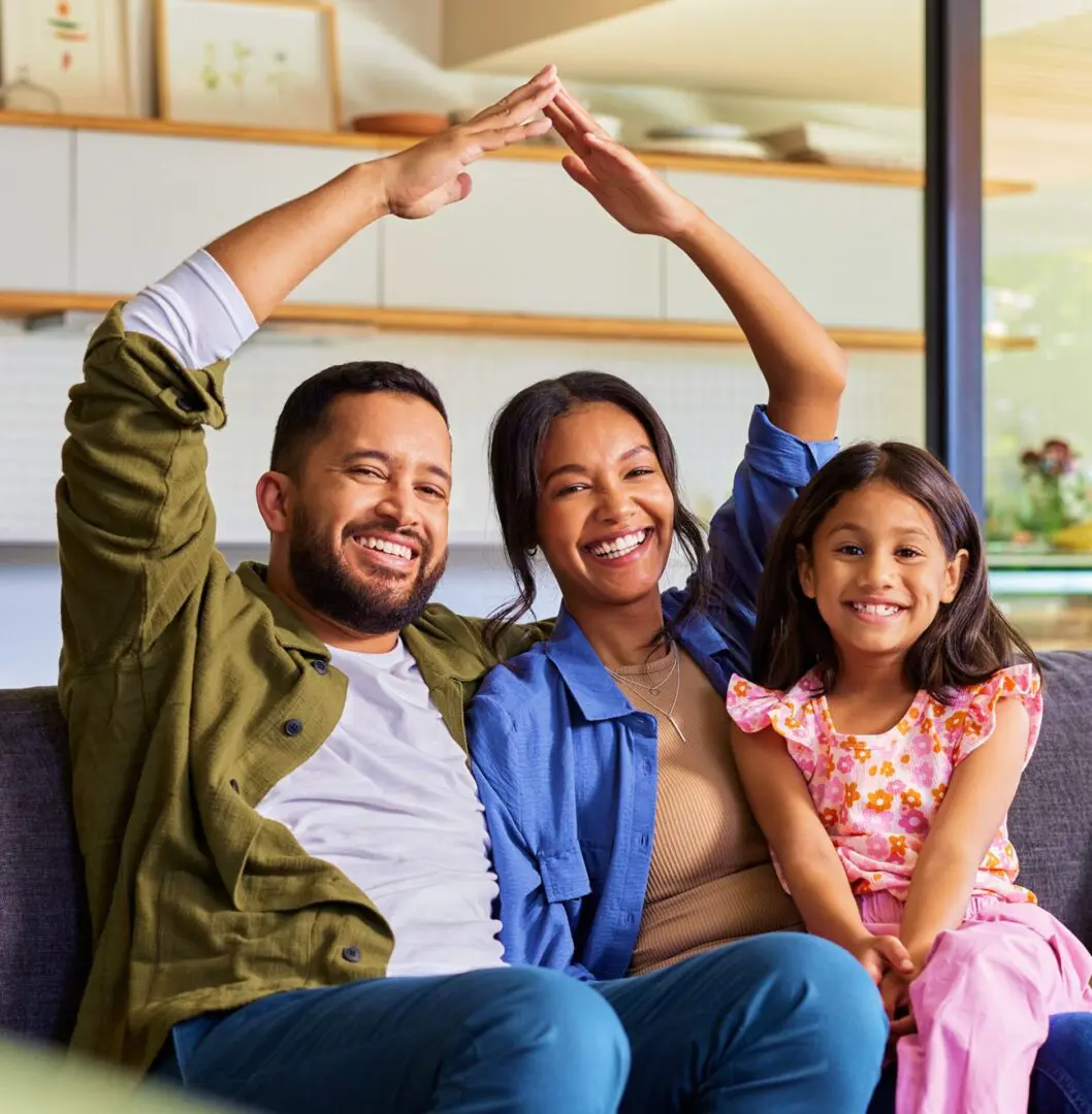 A man, woman, and young girl sit together on a couch, smiling. The man and woman hold their arms above their heads to form a roof shape.