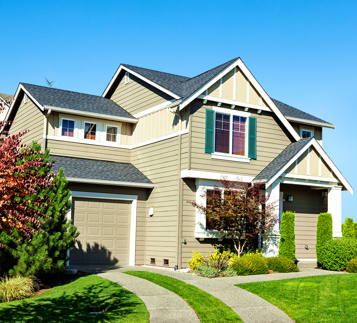 Two-story suburban house with beige siding, green shutters, attached garage, manicured lawn, and driveway under a clear blue sky.